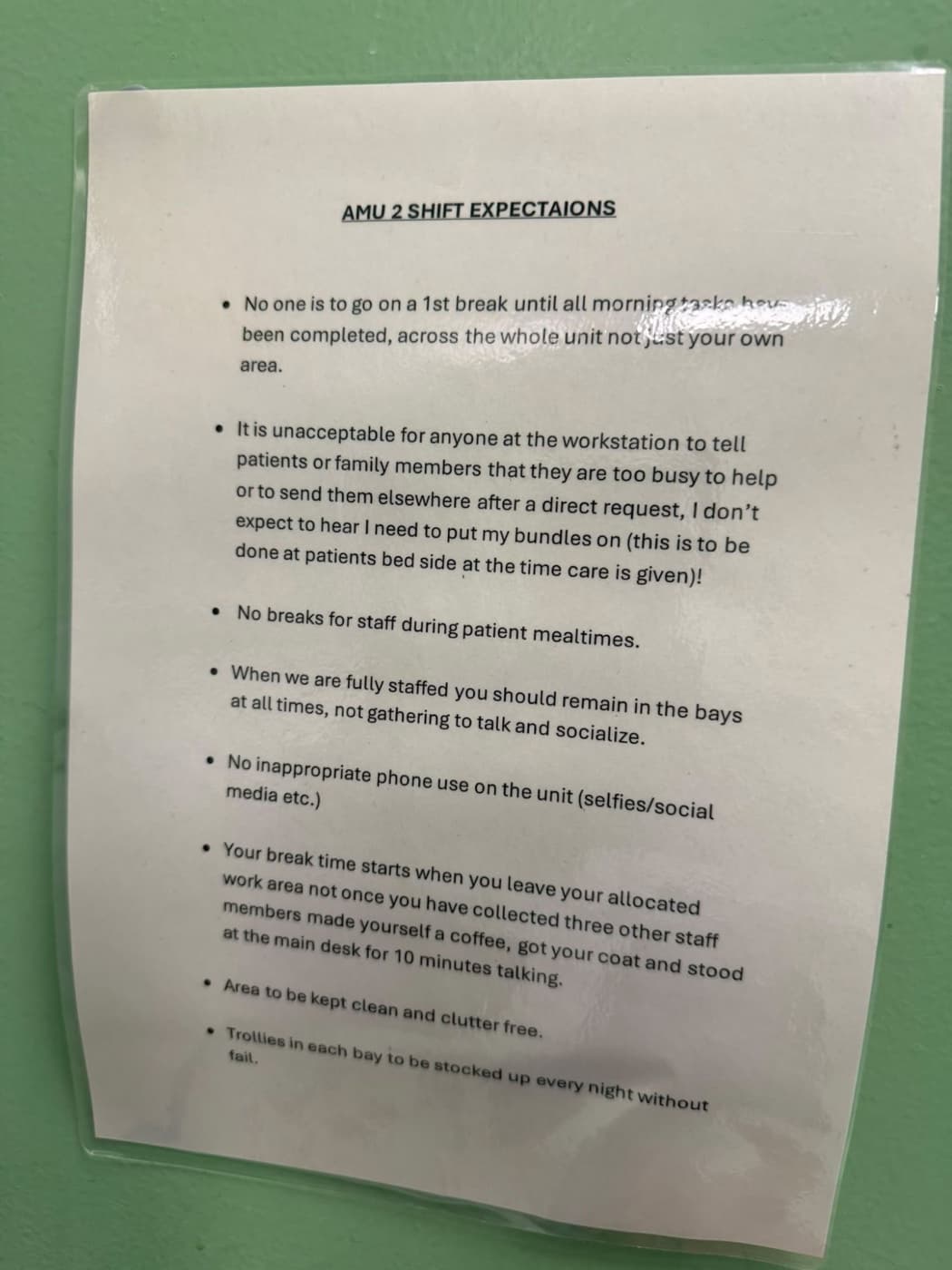 A laminated notice headed AMU 2 Shift Expectations. It lists 8 rules: no one goes on a first break until all morning tasks are done across the whole unit, not just your own area; it is unacceptable to tell patients or family members you are too busy to help or to send them elsewhere, and bundles are to be done at the bedside at the time care is given; no breaks for staff during patient mealtimes; when fully staffed, remain in the bays rather than gathering to talk and socialise; no inappropriate phone use such as selfies or social media; break time starts when you leave your allocated work area, not once you have collected three other staff members, made a coffee, got your coat and stood at the main desk for 10 minutes talking; keep the area clean and clutter free; stock the trolleys in each bay every night without fail.